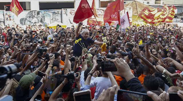 Crowd hoists Lula onto their shoulders, holding flowers and taking picture. Lula has a defiant fist raised.