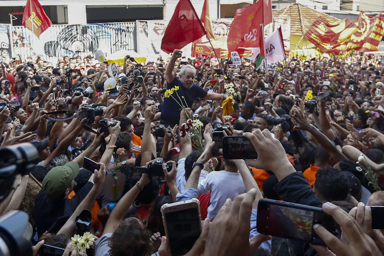 Crowd hoists Lula onto their shoulders, holding flowers and taking picture. Lula has a defiant fist raised.