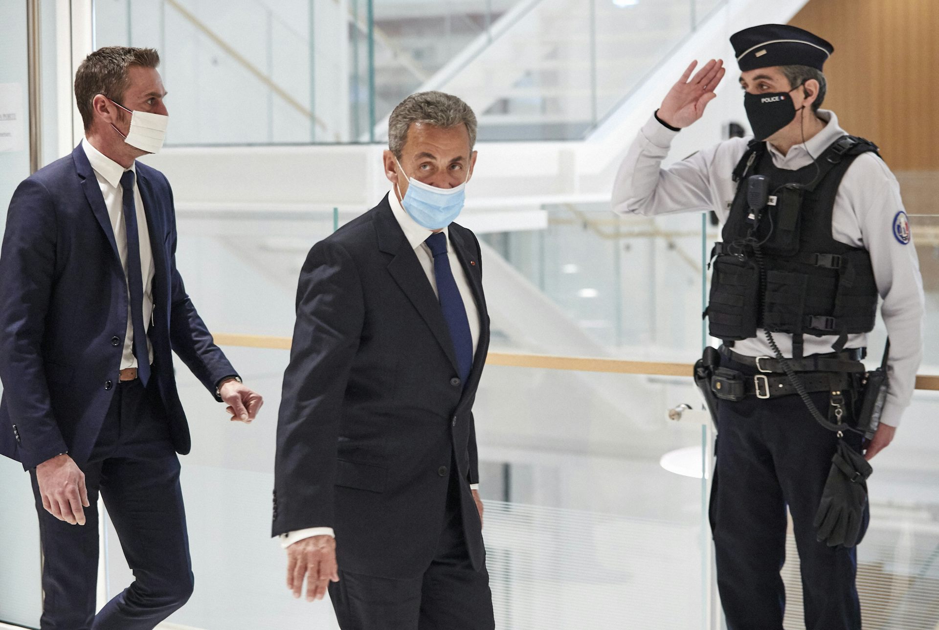 Sarkozy, wearing a face mask, walks through a glass building, trailed by another man in a suit. A police officer salutes.