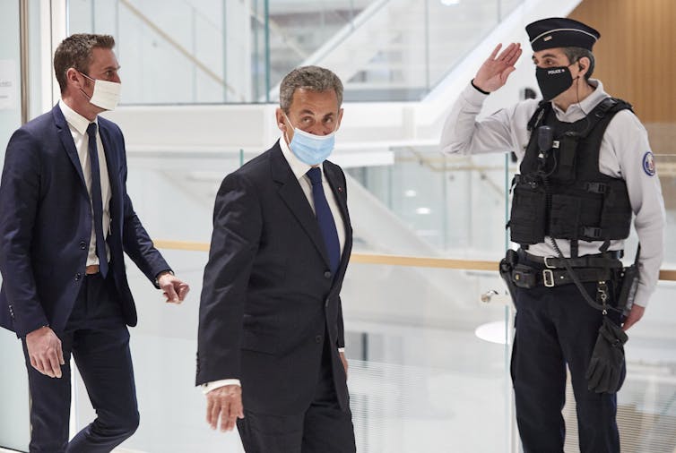 Sarkozy, portant un masque facial, traverse un bâtiment en verre, suivi par un autre homme en costume.  Un policier salue.