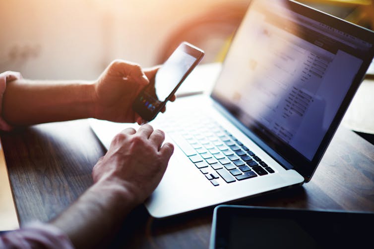 Man's hands holding mobile phone in front of open laptop.