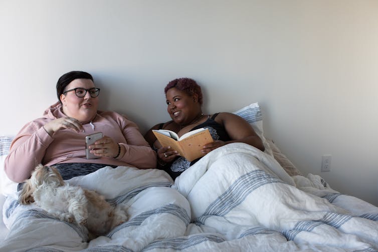 Two women in bed reading book and phone with dog