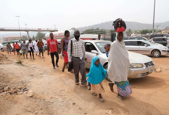 A woman holding a child and carefully balancing a baby on her back and a load on her head, leads a group of people walking on the road side in Abuja, Nigeria's capital city. 