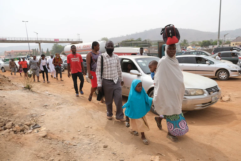A woman holding a child and carefully balancing a baby on her back and a load on her head, leads a group of people walking on the road side in Abuja, Nigeria's capital city.