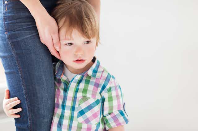 Little boy wearing colourful shirt holds onto woman's jeans