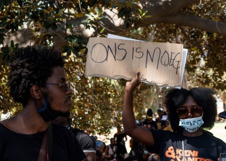 A woman wearing a white COVID-19 mask raises a protest placard while a man looks on.