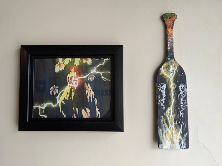 A framed drawing next to a paddle hung on a wall.