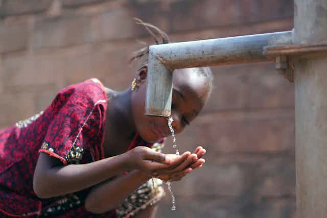 A young girl drinking water from an outdoor pump.