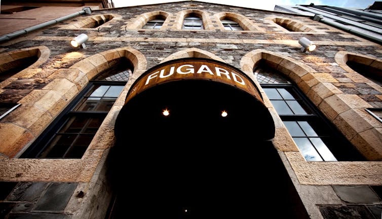 An entrance to a venue, a metal sign reading 'Fugard', the camera pointing up from street level revealing a church-like old stone structure a few storeys high.