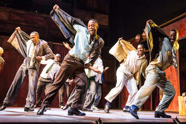 On stage, half a dozen men dance, one arm pulling up their jackets in unison, smartly dressed in 1950s city attire, smiling as they move.