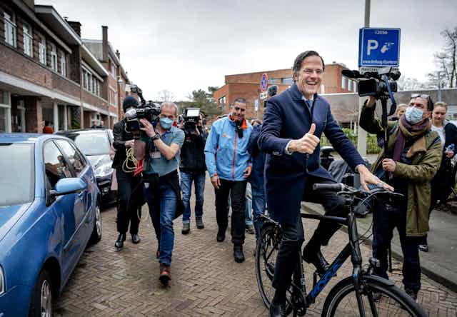 Dutch prime minister cycling down a street as press photographers follow him.
