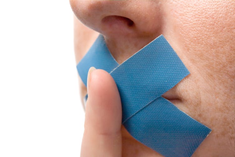 Person with blue tape over their mouths holds finger to lips