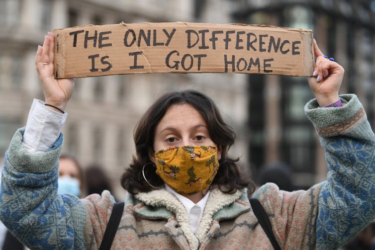 A woman holding a sign reading 'The only difference is, I got home.'