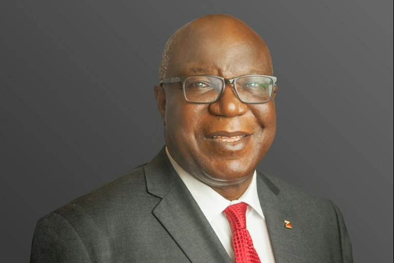 An elder man smiles at the camera. He is balding, with glasses on and a suit with a red tie.