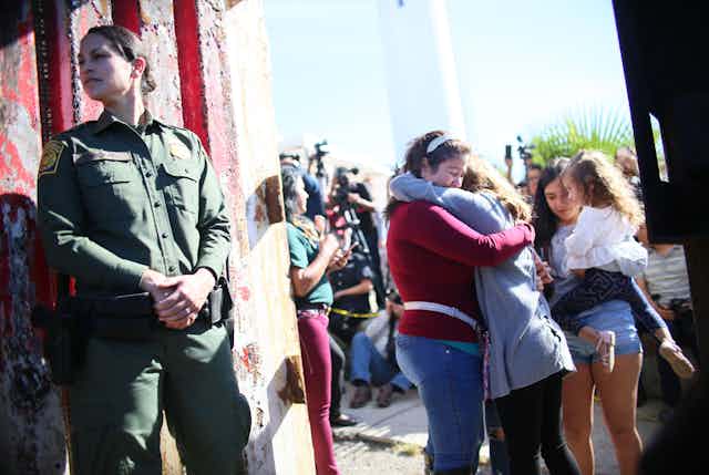 Border patrol guard looks over her shoulder next to border fence as women hug in the background