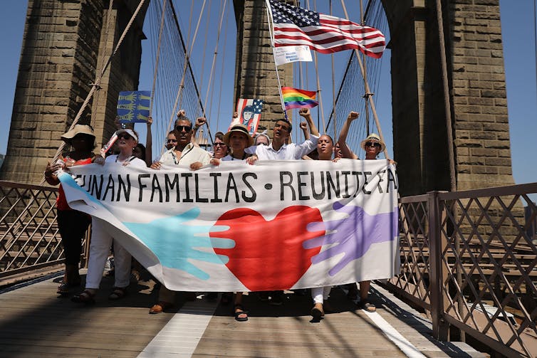 Group crosses the Brooklyn Bridge holding signs that promote 'Reunited Families' and an American flag