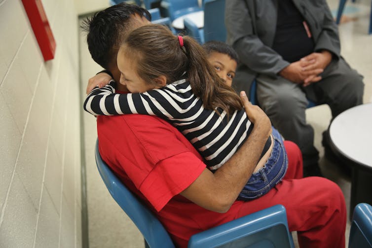 Small girl in striped shirt hugs a man in a red jumpsuit, in an institutional setting