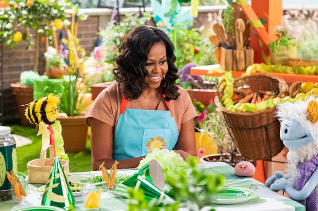 Michelle Obama sits at a table in a garden with puppets.