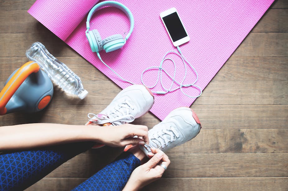 Woman lacing up her running shoes next to her headphones, smartphone, water bottle, kettlebell, and yoga mat.