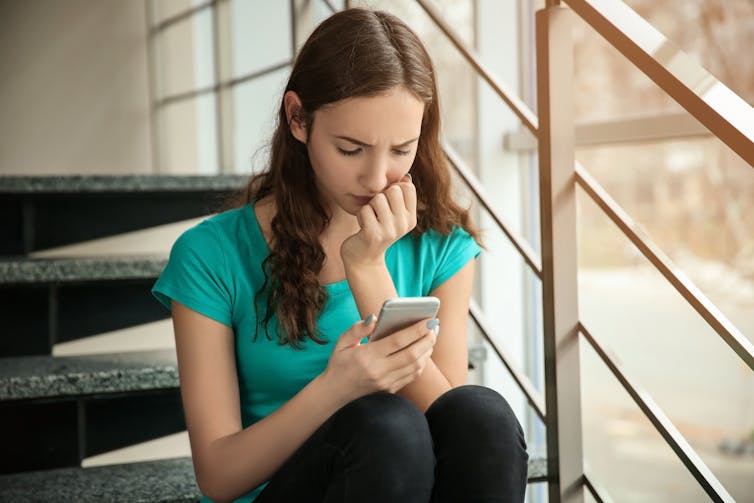 Teenage girl sitting alone looking at her smartphone while looking worried.