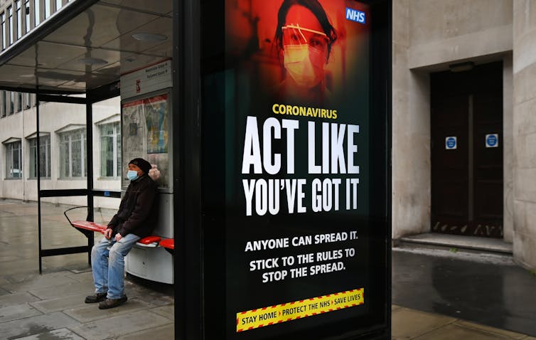 A person waits at a bus shelter with a poster warning about COVID-19.