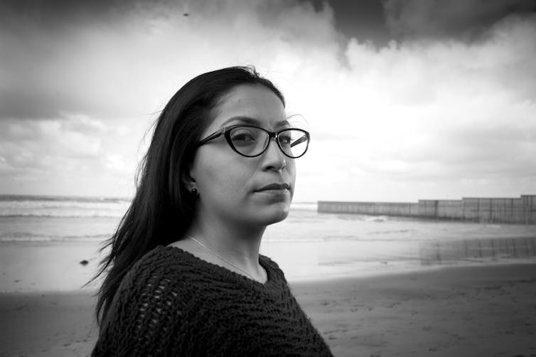 Black-and-white image of a woman standing on a beach with a large fence in the background