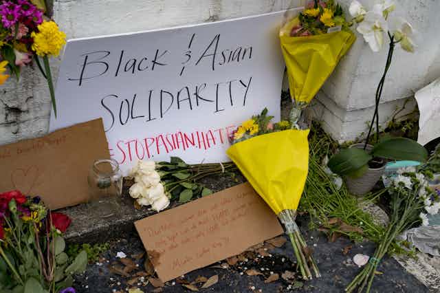 Flowers and a sign reading 'Black & Asian Solidarity STOP ASIAN HATE' lean on a white wall