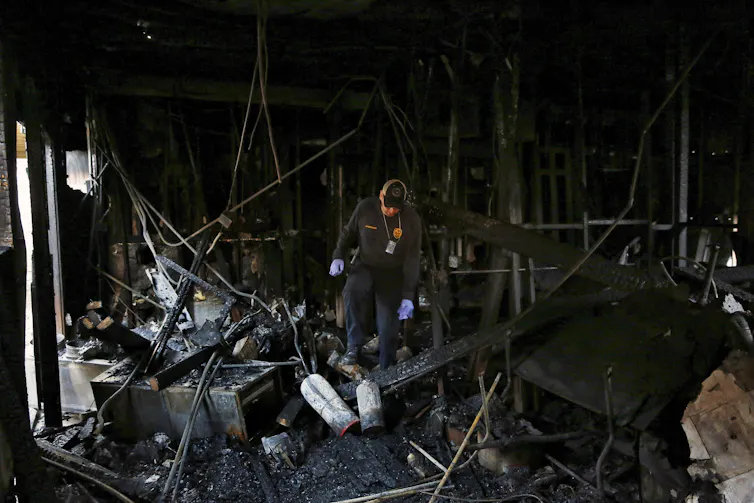 Fire inspector walks through ruins of a charred building, looking at the ground