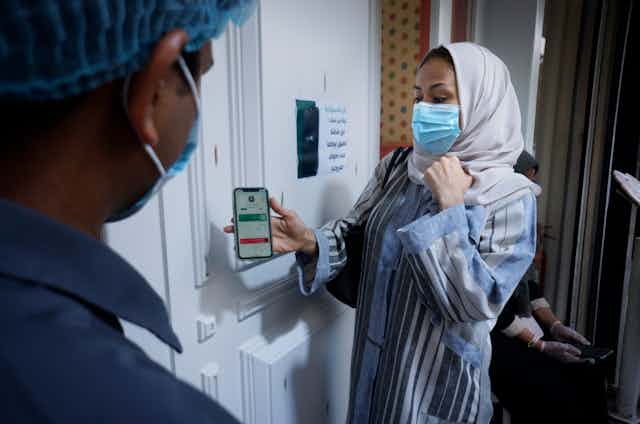 A woman wearing a headscarf and a surgical mask shows a man her smartphone screen