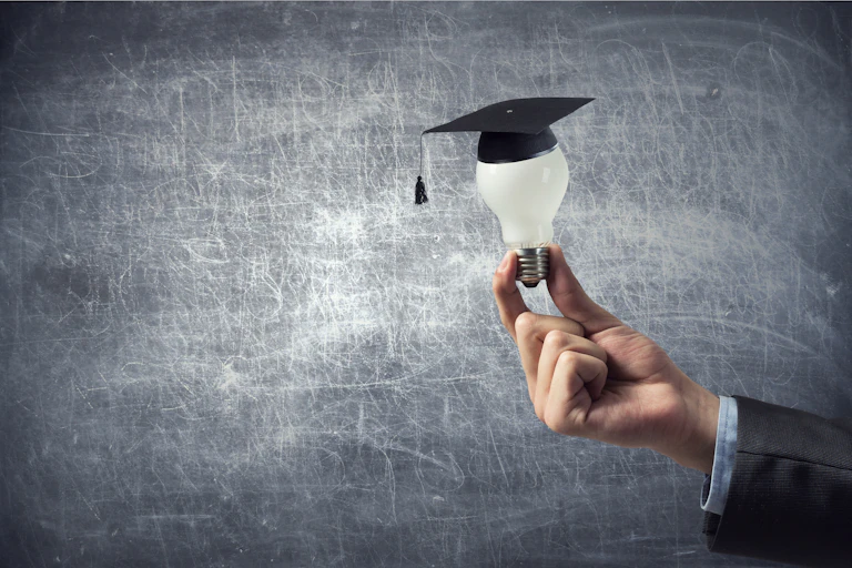 A hand holding a light bulb that has an academic mortarboard hat on the bulb