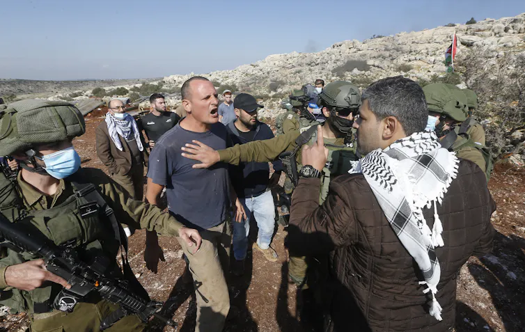 A Palestinian man and an Israeli man argue, with armed Israeli soldiers looking on.