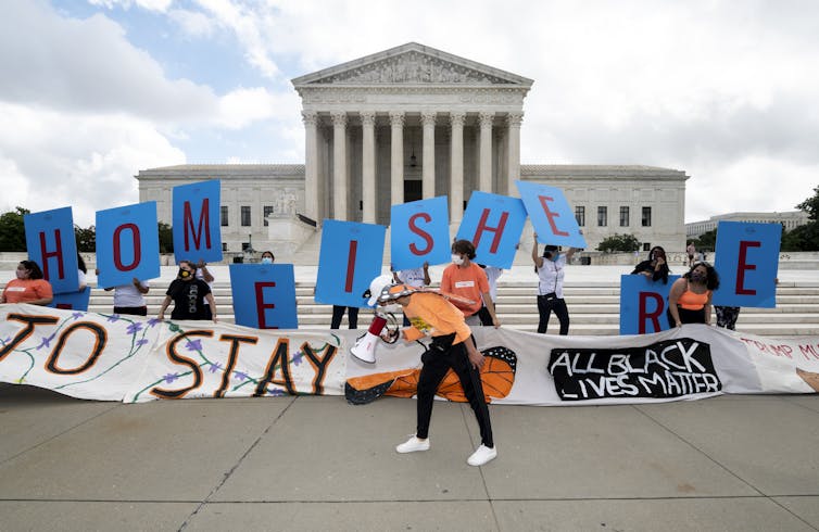 Group of young people hold up signs rendering 'Home is Here' and 'Here to Stay' with Supreme Court in background