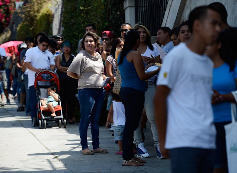 Line of several dozen young adults, some with young children, standing in the sun