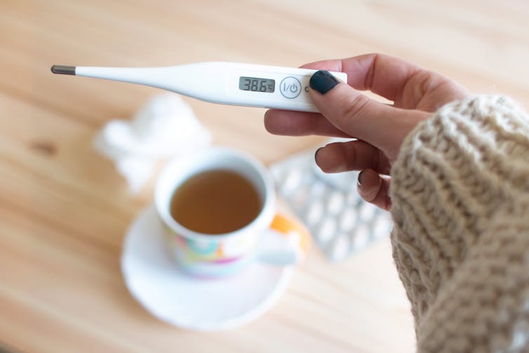 A hand holds a thermometer. There is a cup of tea and tablets on a table in the background.