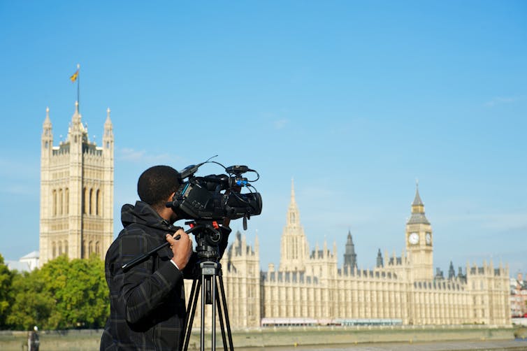 A man with a TV camera filming across the River Thames to Westminster Palace and the Houses of Parliament.
