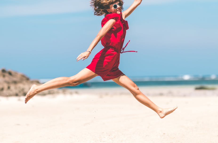 Woman in red jumpsuit jumping at the beach.