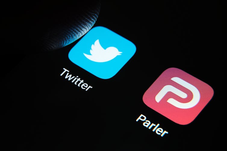 A thumb hovers over a phone screen featuring the icons for Twitter and Parler