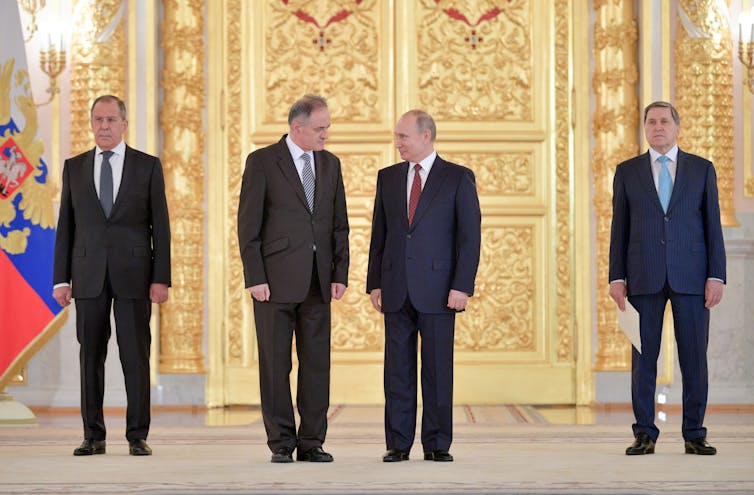 Putin and another man in a suit look at each other intensely, flanked by two other serious-looking men, against a gilded backdrop