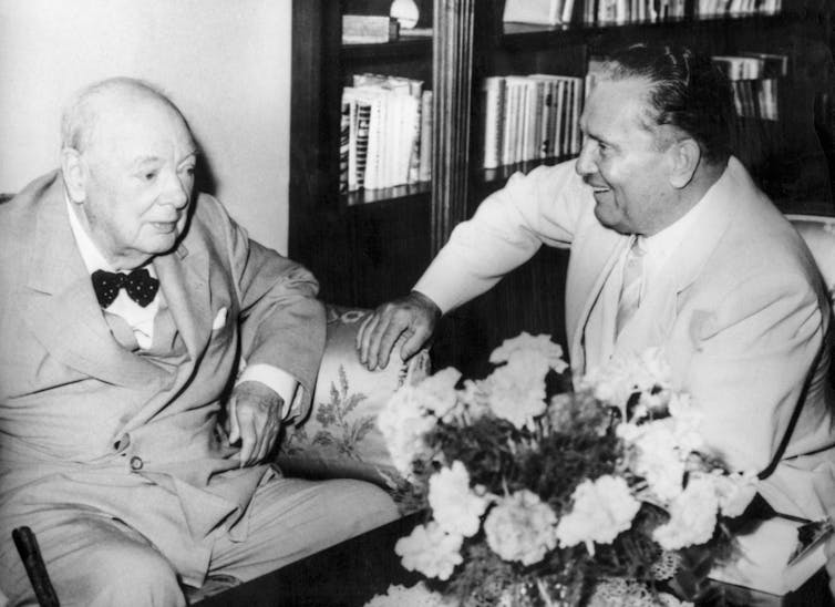 Black-and-white image of a very elderly Churchill sitting and laughing with a younger Tito