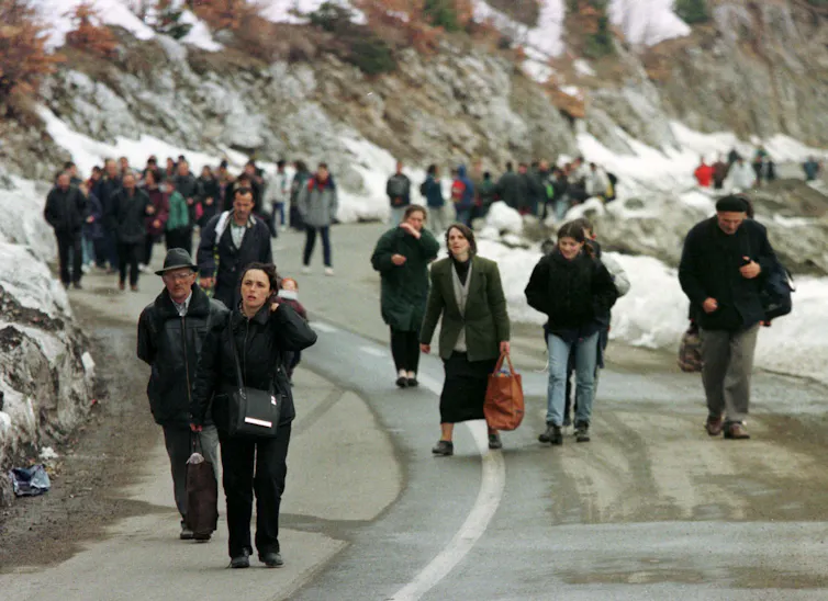 People walk on a mountainous road, wearing backpacks and carrying language; there is snow on the ground