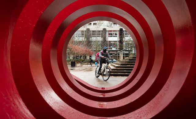 A food delivery worker wearing a face mask is framed by the red circles of a large public art installation. 