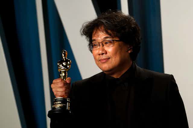 Korean man dressed in black holds Oscar trophy.