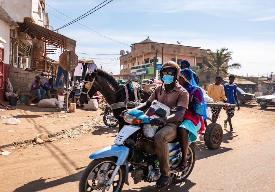 A street scene in Mbour, Senegal featuring a man and a woman on a scooter and a horse-drawn cart. 
