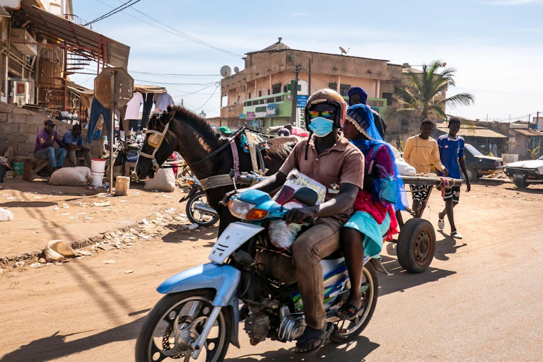 A street scene in Mbour, Senegal featuring a man and a woman on a scooter and a horse-drawn cart.