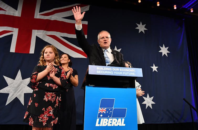 Scott Morrison, with his family beside him, holds up his hand behind a Liberal party podium.