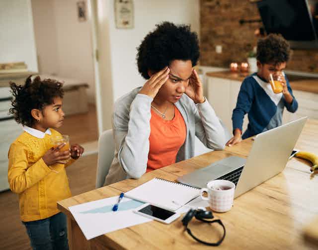 A Black woman in front of her laptop looking stressed as two children drink juice