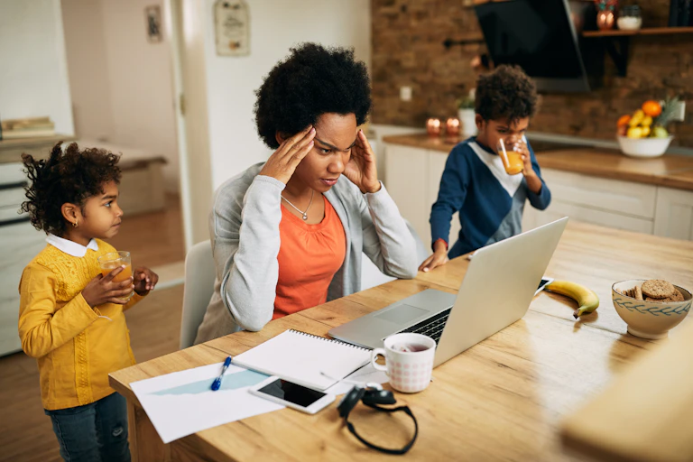 A Black woman in front of her laptop looking stressed as two children drink juice