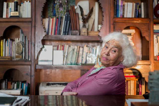 In study full of carved bookshelves, books and trinkets, an elder woman with grey hair and a dirty pink top leans back and looks into camera, her mouth in a pout and her eyes challenging.