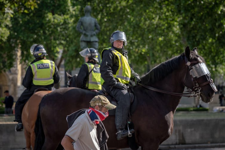 Police on horseback with helmets as a masked passerby in army cap walks past