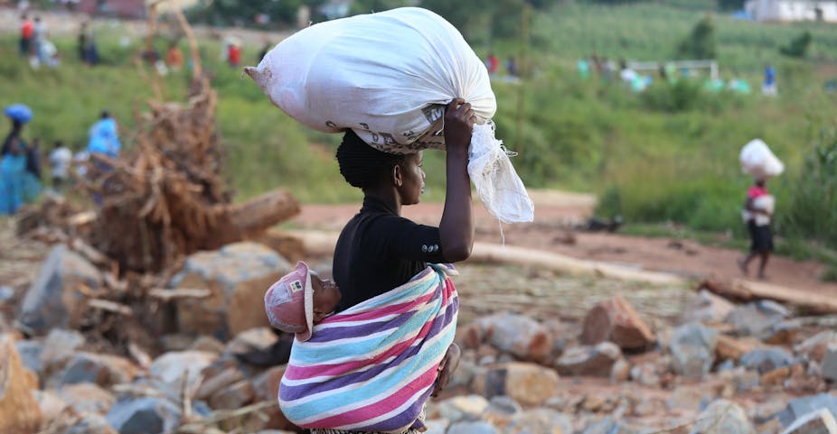 Woman carries a white sack on her head and a baby on the back, wrapped in a blanket.
